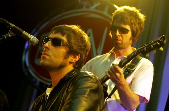 Oasis perform on stage in 90s. Liam Gallagher is in foreground singing while Noel Gallagher plays guitar behind him.