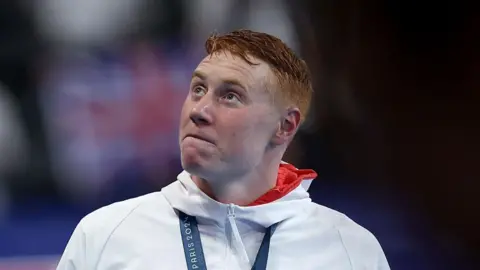 Getty Images Tom Dean, who has red hair and wears a white and red hoodie with a medal around his neck, looks to the side, with a Union Flag seen in the background 