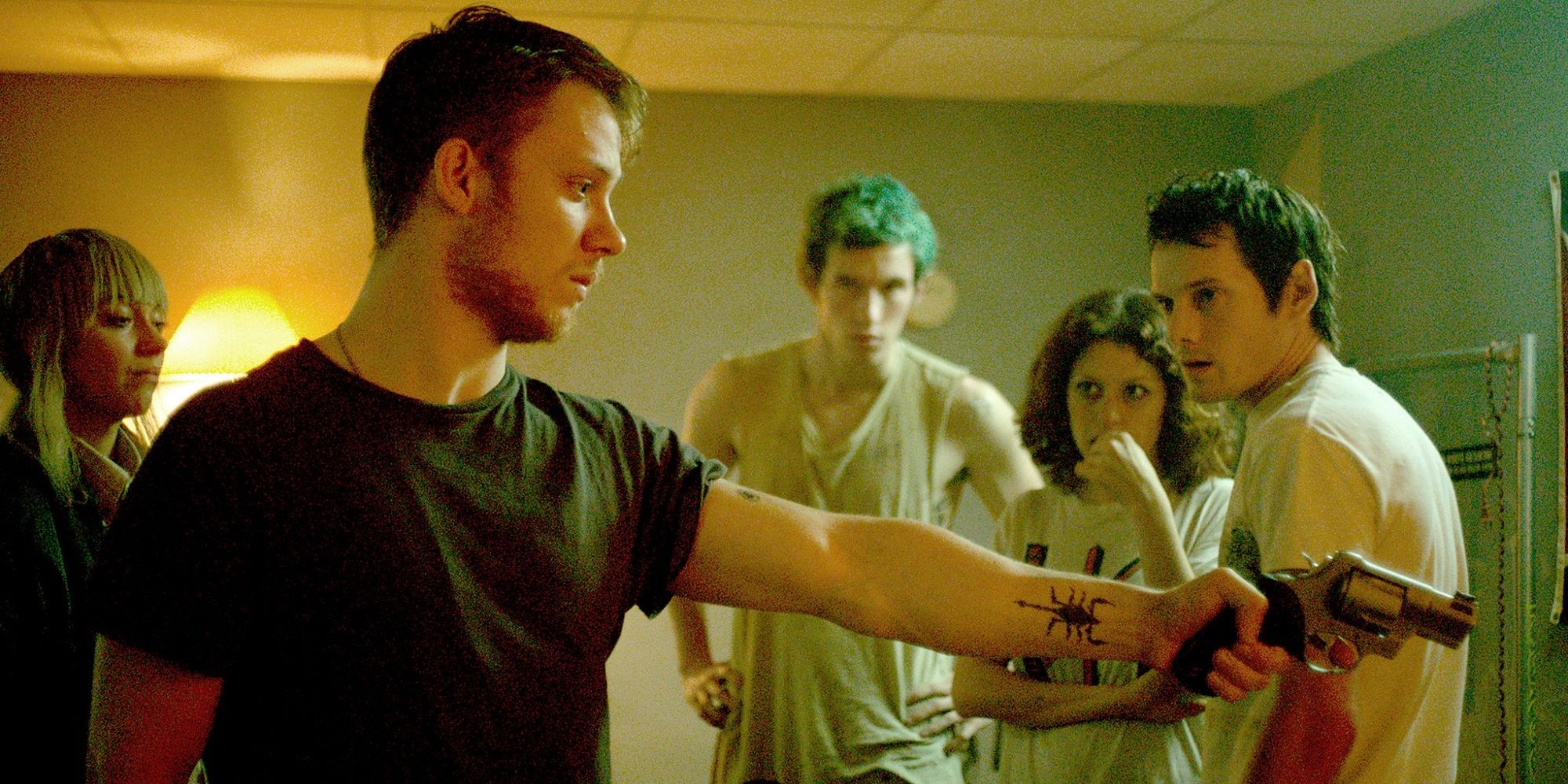 Joe Cole's character, Reece, pointing a gun at someone off screen in 'Green Room' while Anton Yelchin, Imogeen Poots, Callum Tanner, and Alia Shawkat watch on.