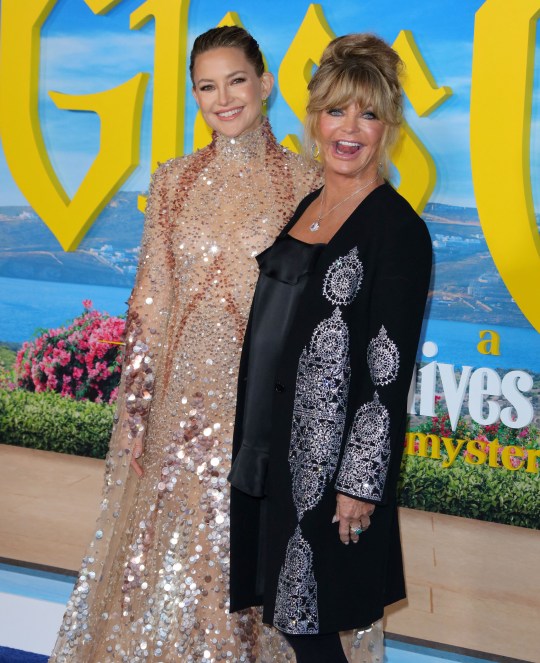 Kate Hudson and Goldie Hawn smiling as they pose together on the red carpet. Kate is wearing a sequinned beige gown and Goldie wears a black outfit