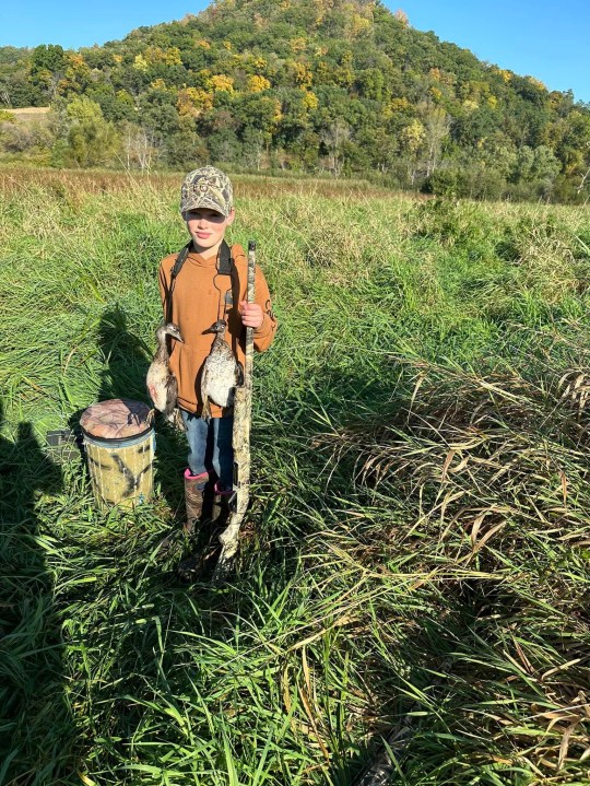 The boy, Owen Beireman, holds a rifle and two shot down birds hung around his neck in a grassy area