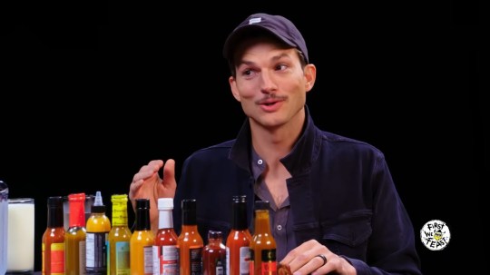 Ashton Kutcher talking on Hot Ones, with various hot sauce bottles in front of him