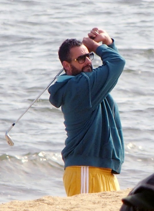 Adam Sandler on the set of Happy Gilmore 2 raising a golf club above his head. He is wearing a blue hoodie and yellow jogging bottoms