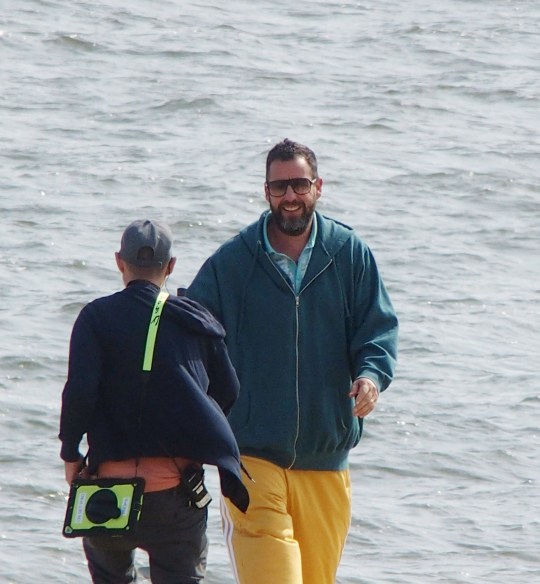 Adam Sandler on the set of Happy Gilmore 2 speaking to a crew member. He is wearing a blue hoodie and yellow jogging bottoms