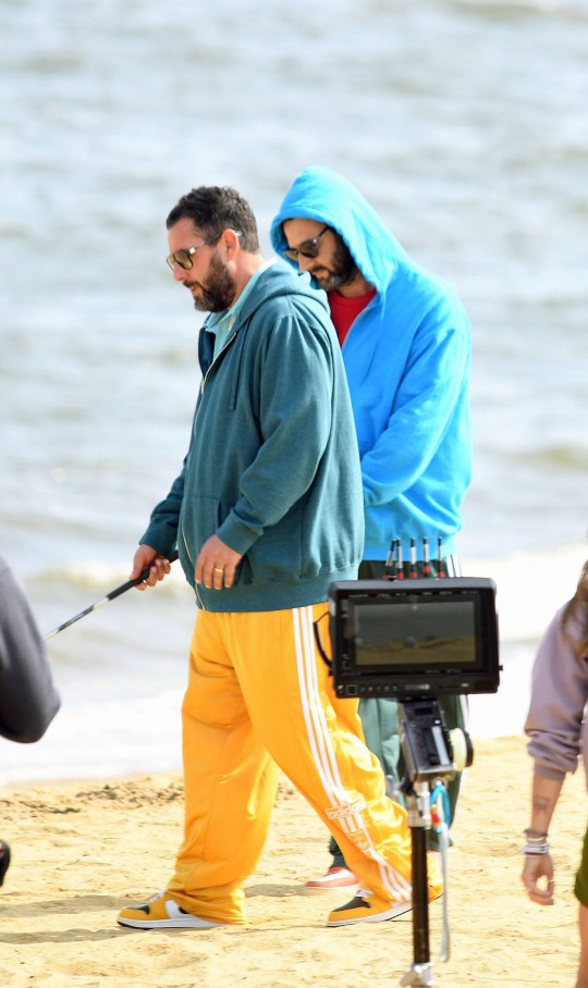 Adam Sandler on the set of Happy Gilmore 2 holding a golf club. He is wearing a blue hoodie and yellow jogging bottoms