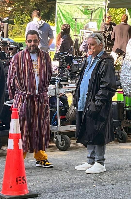 Adam Sandler on the set of Happy Gilmore 2. He is wearing a red striped bathrobe and black sunglasses. He is standing next to a man wearing a black coat, blue shirt, and grey jeans