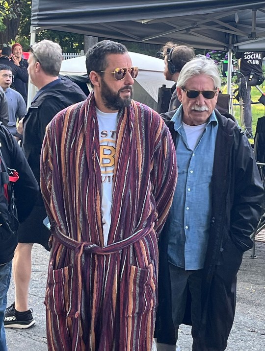 Adam Sandler on the set of Happy Gilmore 2. He is wearing a red striped bathrobe and black sunglasses. He is standing next to a man wearing a black coat, blue shirt, and grey jeans