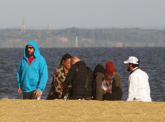 Adam Sandler and crew members on the set of Happy Gilmore 2. He is standing on a beach and wearing a blue hoodie with grey jogging bottoms