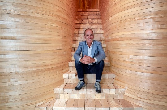 Kevin McCloud sitting on a wooden staircase in Grand Designs. He is wearing jeans, with a white shirt and blazer.