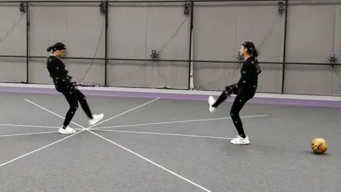 Sports Interactive Two women wearing black, skin-tight motion capture suits with white, ping-pong ball-style spheres attached to them, make kicking motions like those you'd see in a game of football. Numerous small cameras are attached to the grey walls of the studio to record their movements from different angles.