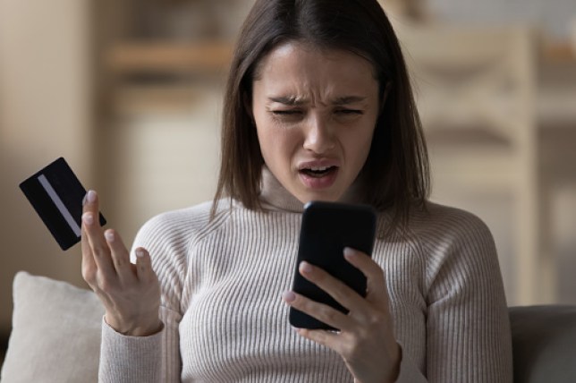 A woman looking frustrated at her phone holding a credit card 