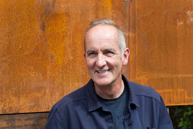 Kevin McCloud smiling. He is wearing a blue shirt and standing in front of a wooden build