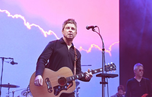 Noel Gallagher on stage with guitar in hand. Background is purple as he looks out to crowd and sings into microphone.