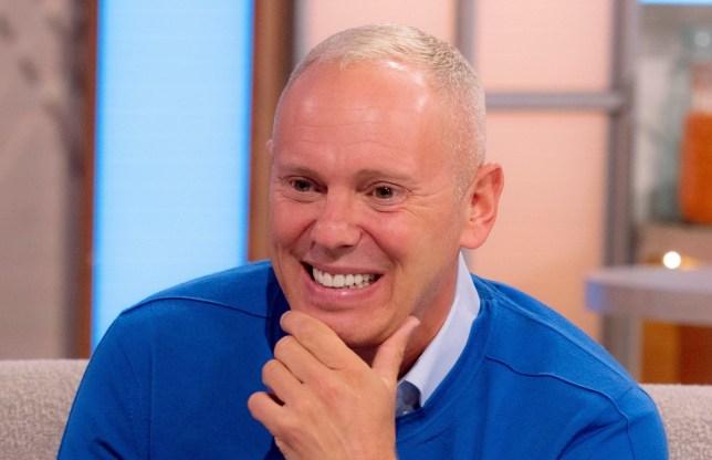 Rob Rinder wearing a blue sweater and a blue collared shirt, smiling with a hand resting on his chin