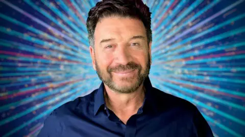 BBC Nick Knowles, a man with dark hair and stubble, wears a dark blue shirt and stands against a blue starburst background, smiling 