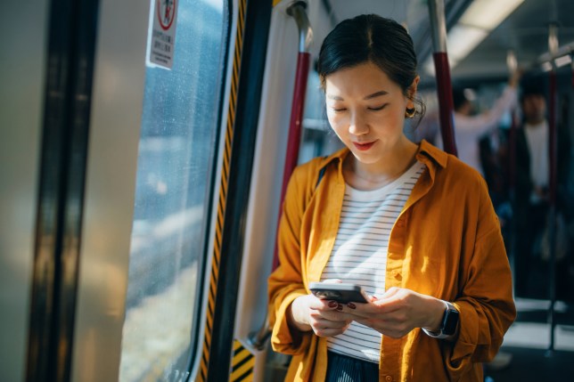 Young woman standing by the window against sunlight in subway train while using her smartphone. 