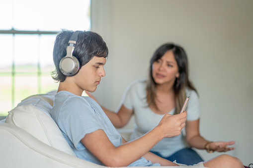 A teenage boy wearing headphones stares at his phone while a woman looks exasperated in the background.