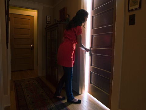 A woman peers through a door slightly ajar from a dark hallway. Light streams through a chink in the door