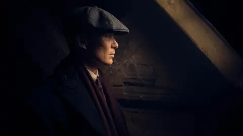 BBC/Caryn Mandabach Productions Ltd./Matt Squire The actor Cillian Murphy dressed as Tommy Shelby, the character he plays in the drama Peaky Blinders. He is wearing a black and white tweed flat cap, black jacket, white shirt, and maroon scarf, and a tie. He is standing inside a dark building with wooden beams.