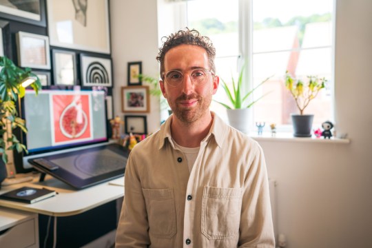 Tom Hovey sitting in a room with a computer and digital sketch pad. He is wearing a cream shirt and glasses while smiling at the camera