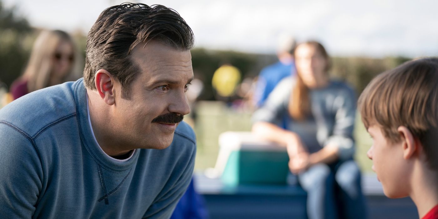 Jason Sudeikis as Ted Lasso leaning down to speak to a young boy at an outdoor sporting event.