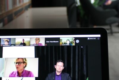 En datorskärm i förgrunden och en suddig person i bakgrunden, som håller i ett webinarie