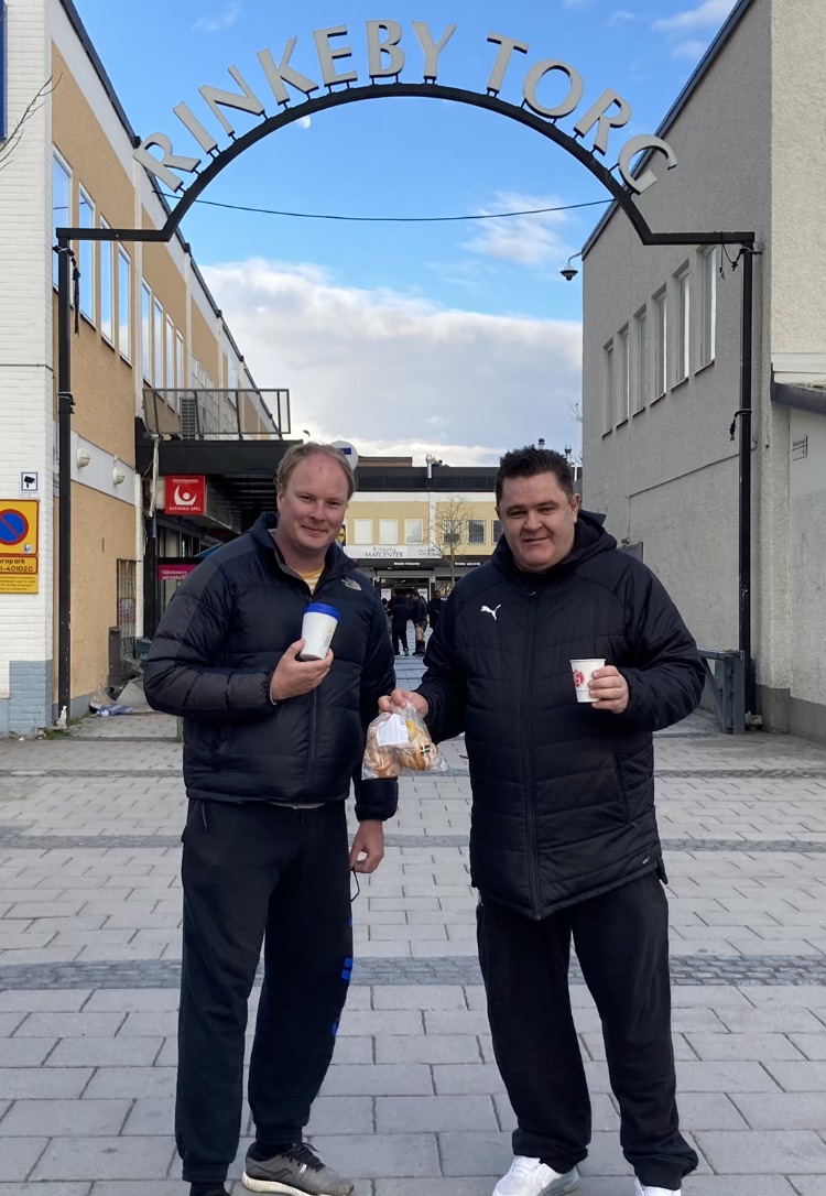 Två män med kaffemuggar i händerna på Rinkeby torg.