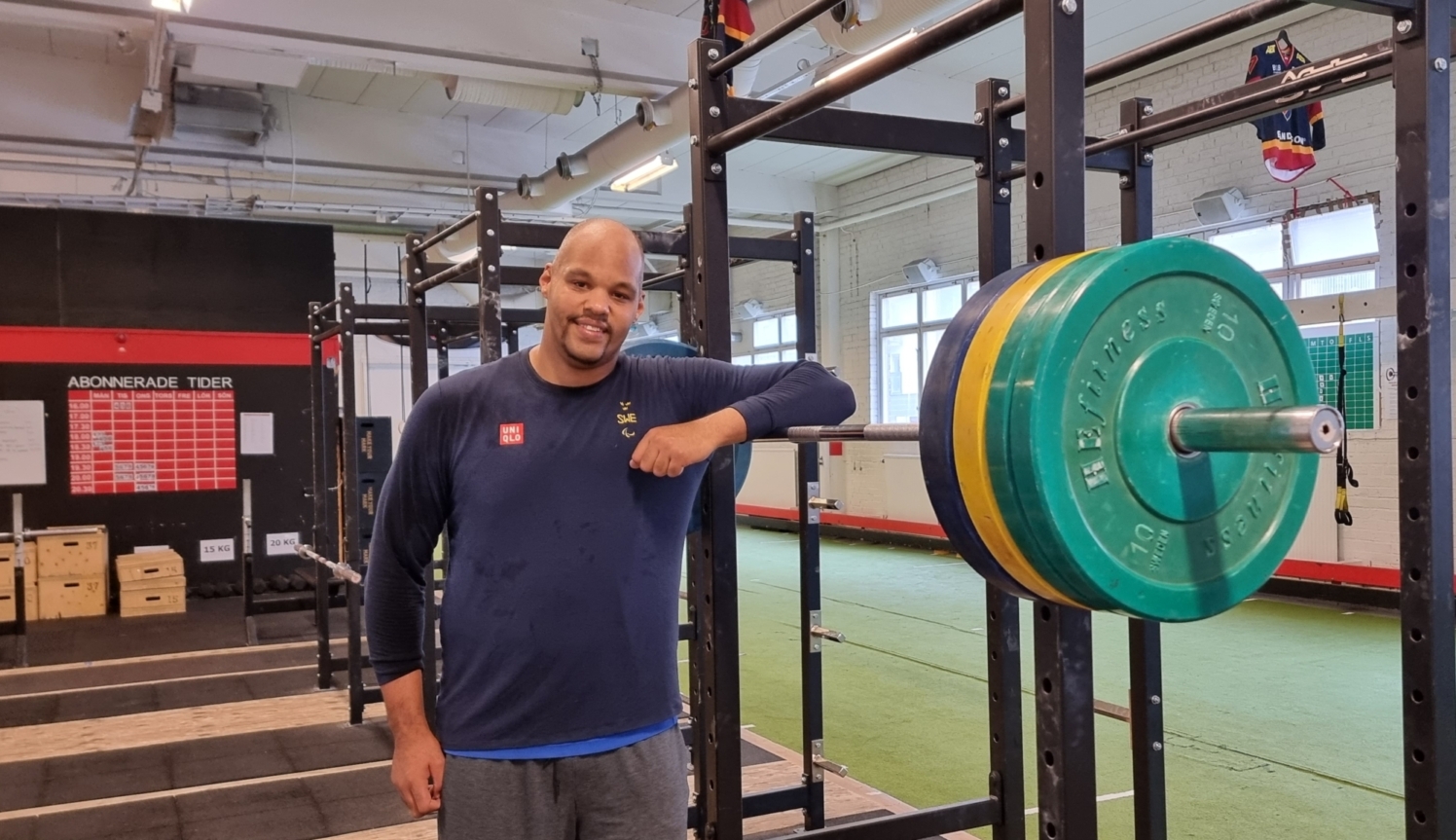 en leende man i ettt gym lutar sig mot en skivstång