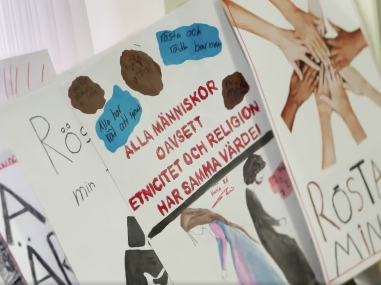 Fyra plakat som ligger omlott. Främst ett plakat med sju händer som möter varandra och texten "Rösta för min skull". Bakom den ett plakat med texten "Alla människor oavsett etnicitet och religion har samma värde!" med två människor målade i vattenfärg.