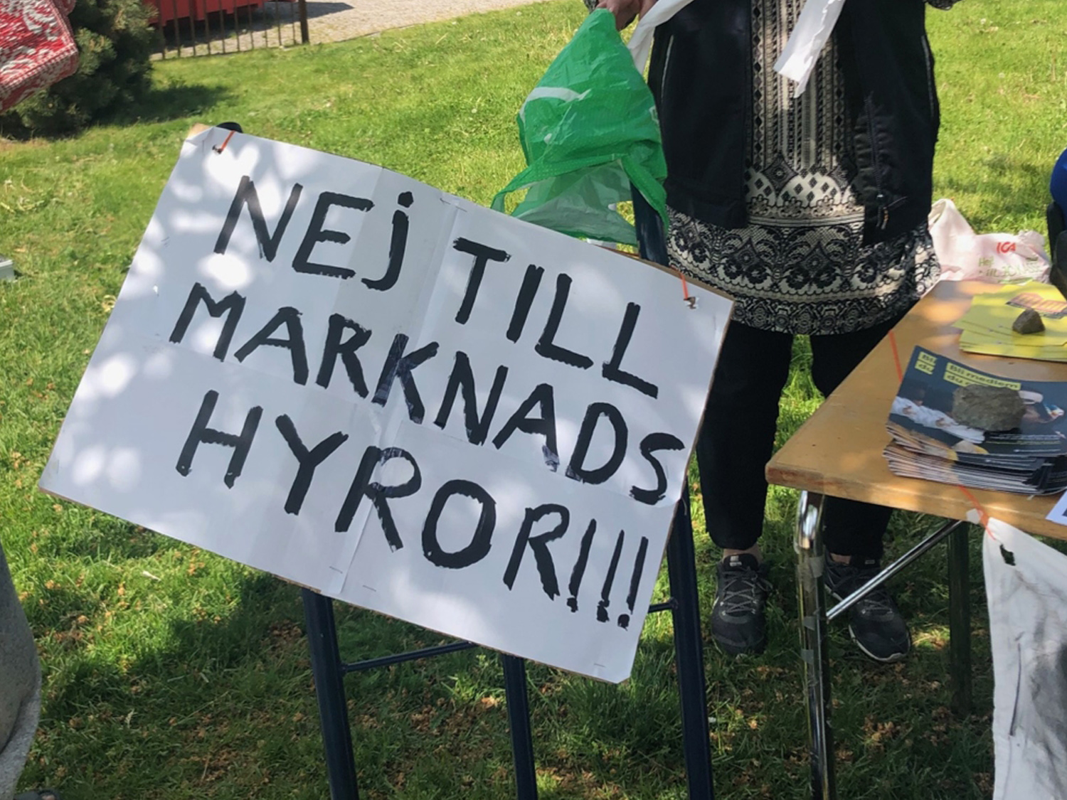 En kartongskylt med texten NEJ TILL MARKNADSHYROR!!! Skylten är upphängd på en pall som står på en grön gräsmatta. Bakom skylten skymtas en kvinna i jympaskor, tunika och jacka.