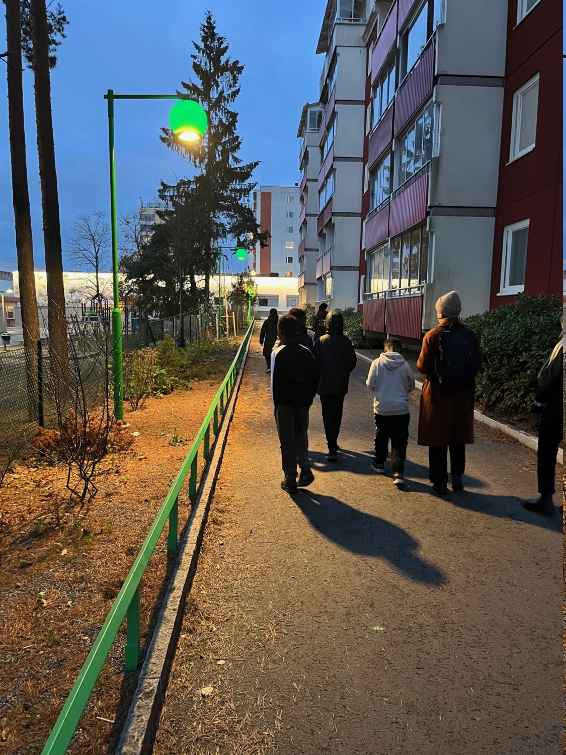 Barn och vuxna går längs en väg i Husby, gröna gatlyktor.