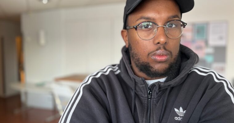 Ung somalisk man i keps och glasögon. Ser bister ut.