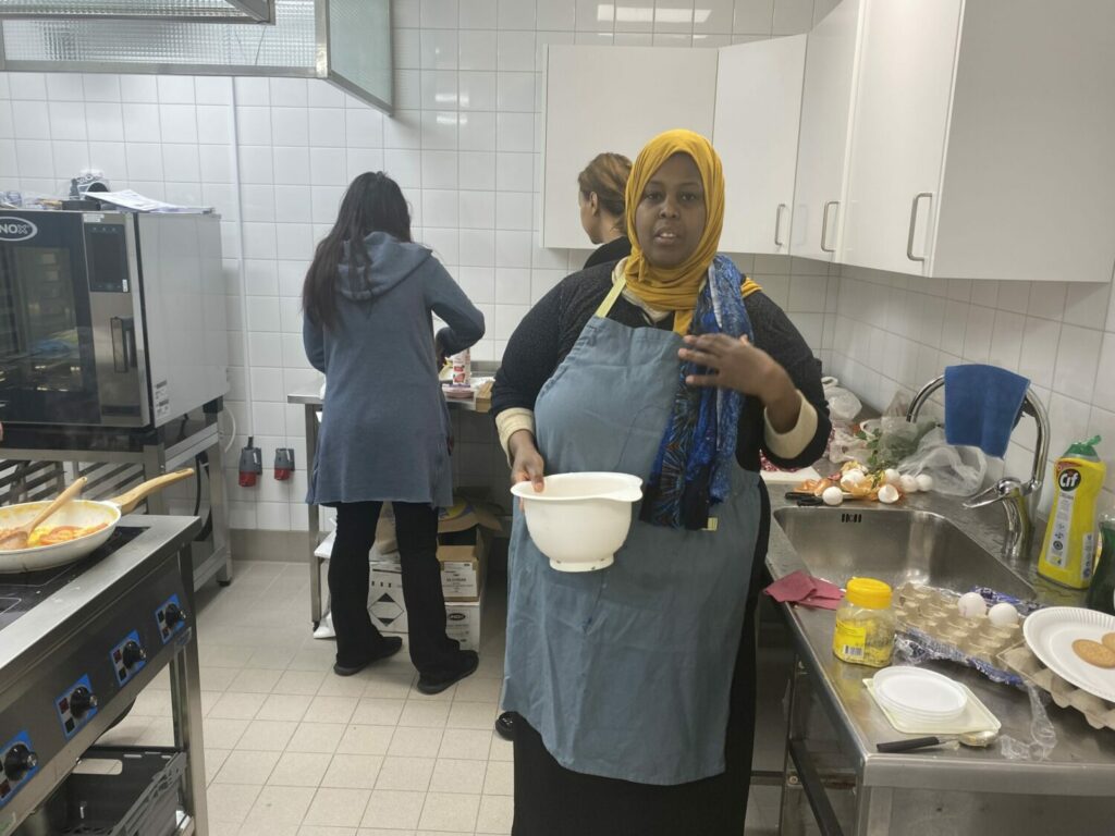 Kvinna i förgrunden med en plastbunke i handen, två kvinnor i bakgrunden arbetar vid köksbänk