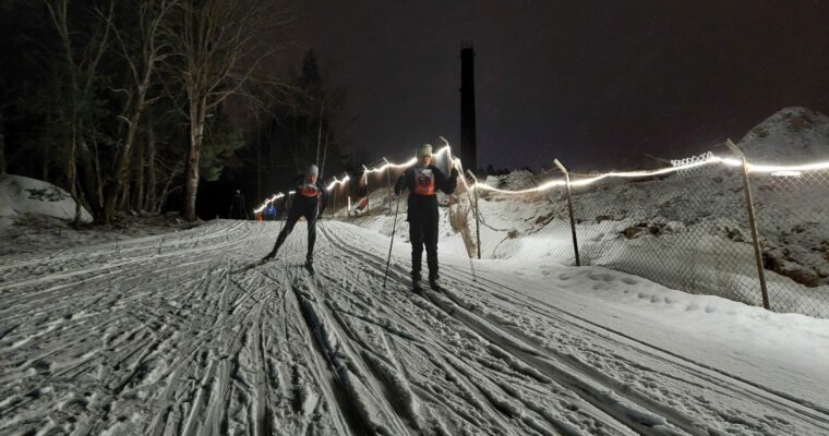 Två skidåkare i skidspåret, kvällstid med lampor på.