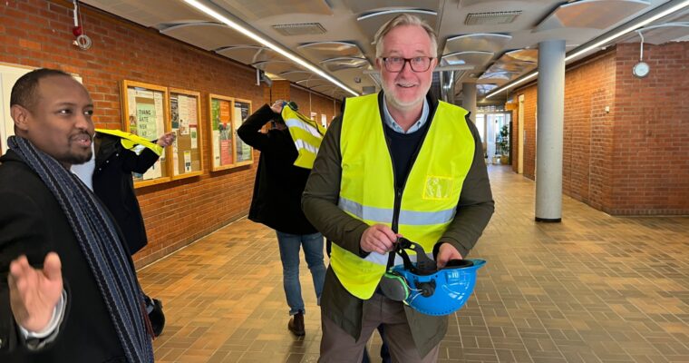 Leende man i gul väst och med en bygghjälm i handen.