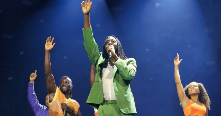 man i grön kostym sträcker upp ena handen tillsammans med dansare på en scen