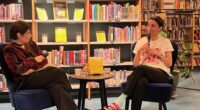 Två kvinnor sitter i ett bibliotek och samtalar på en scen.