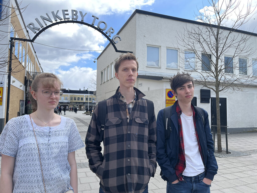 en tjej och två killar ser allvarliga ut framför Rinkeby Torg-skylten
