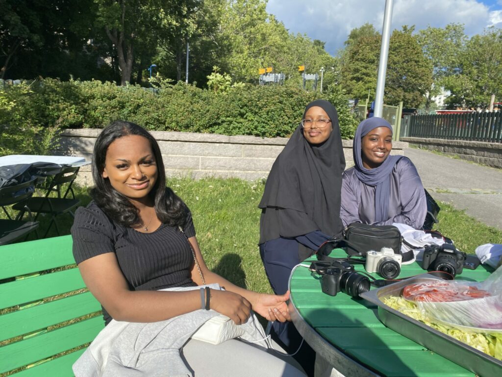 Tre unga kvinnor sitter på en parkbänk och ler mot kameran