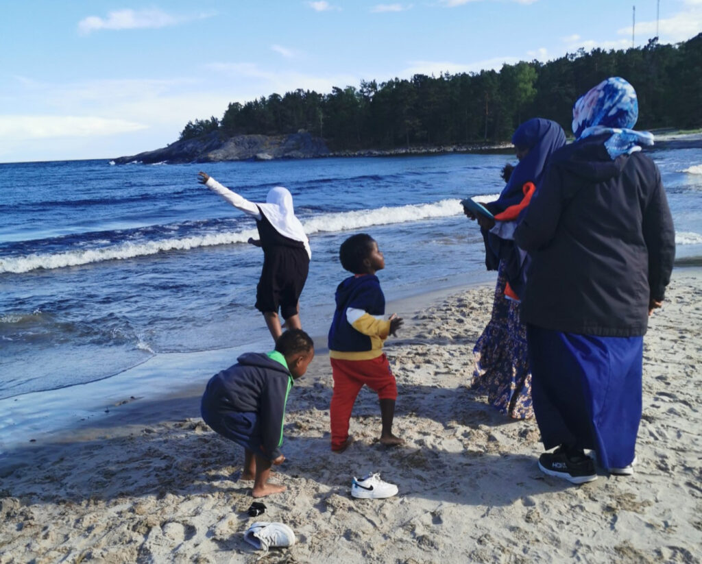 barn doppar fötter i kallt havsvatten vid en strand och två kvinnor tittar på