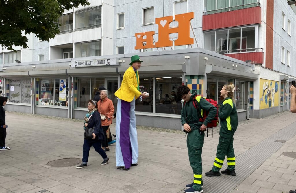En man utklädd till clown går på höga styltor, ambulanspersonal tittar på.