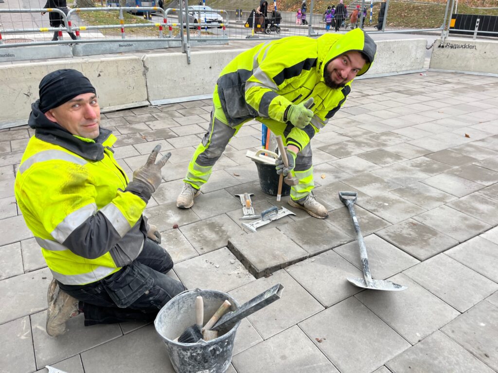 Två arbetare i gula varseljackor som lägger om plattor på trottoaren.