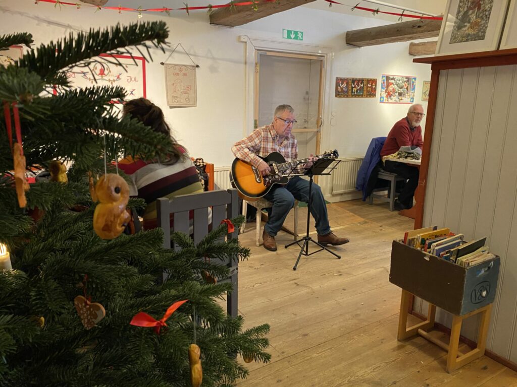 En man sitter på en stol och spelar gitarr och sjunger. En julgran i högra kanten av bilden