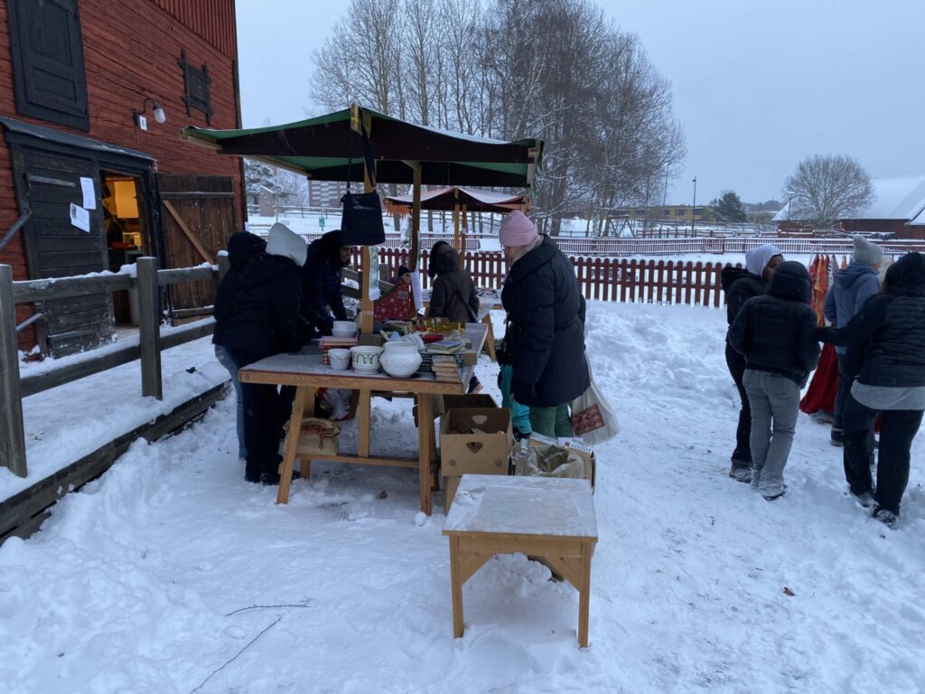 Människor vid ett bord med loppisförsäljning utomhus på snötäckt gårdsplan
