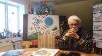 En gråhprig man i sina bästa år, sitter framför sina konstverk och tittar rakti in i kameran.