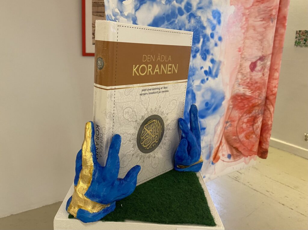 Ett konstverk där två händer av gips målade som svenska flaggan håller i Koranen