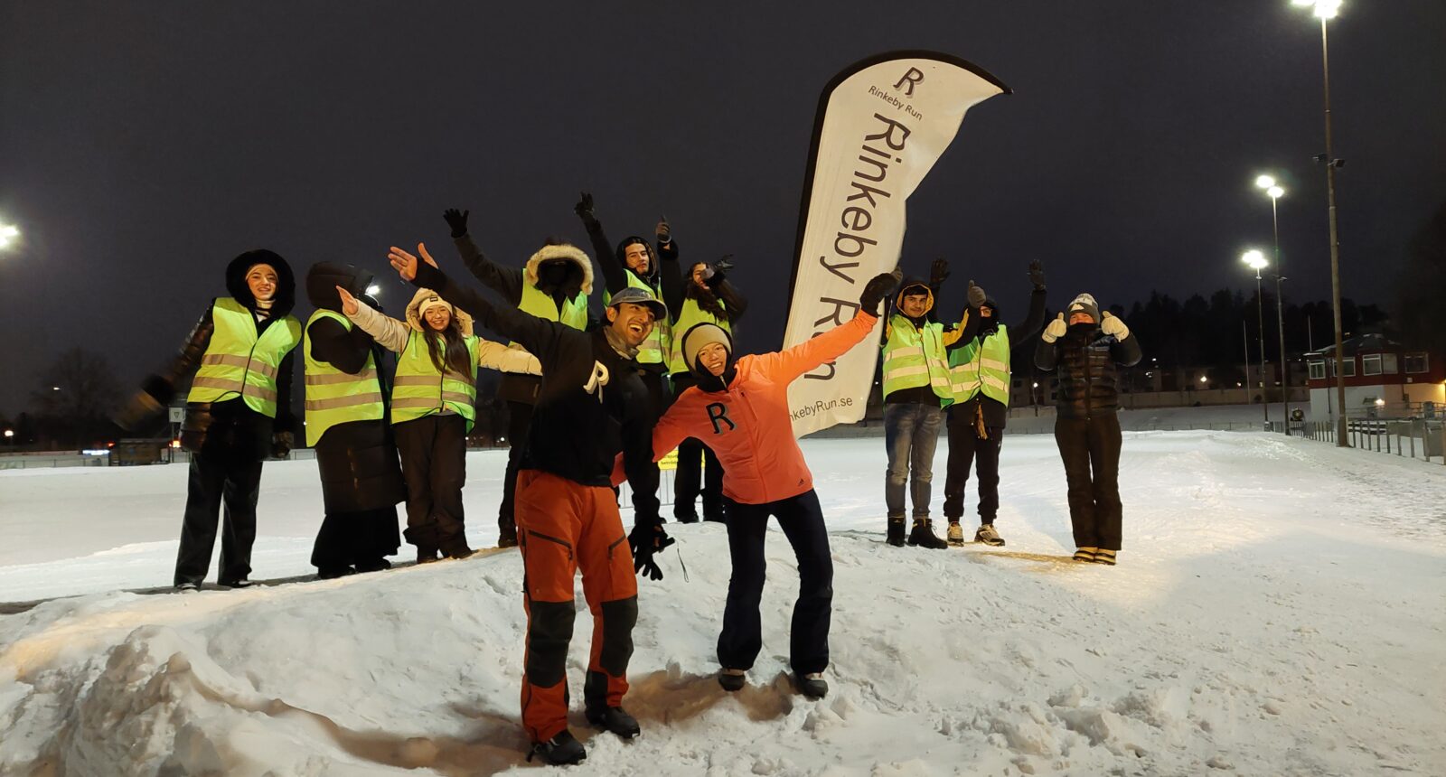 vinterklädda personer står i snödrivor framför idrottsplats
