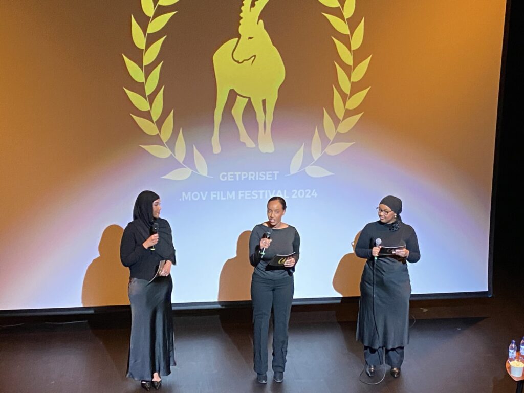 Tre unga kvinnor på en scen framför en filmduk med loggan "Getpriset .MOV Film Festival 2024"
