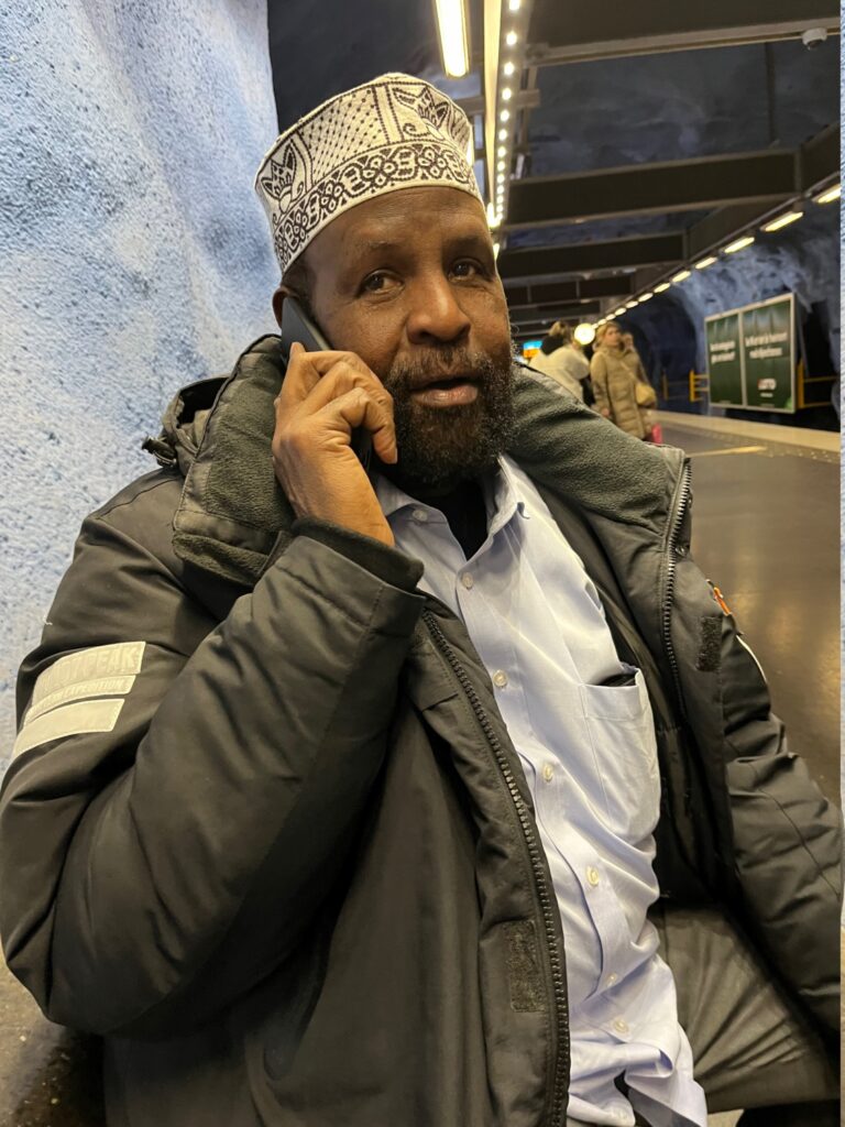 En äldre somalisk man sitter på en bänk i t-banan och talar i en mobil.