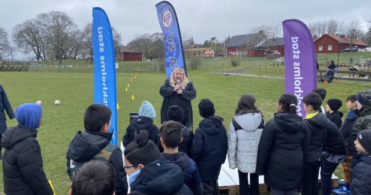 Kvinna talar på en scen utomhus, barn står och lyssnar